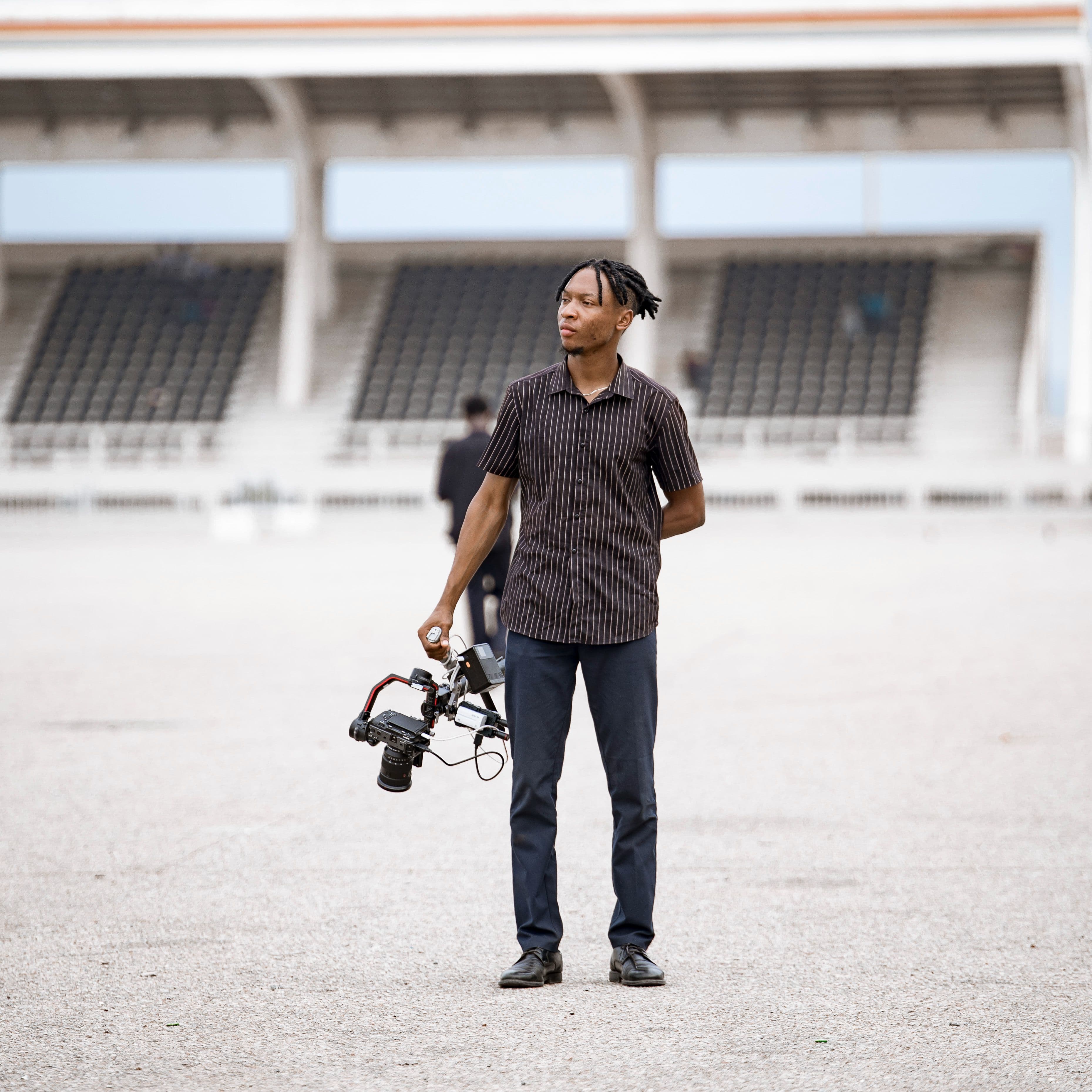 The filmmaker standing alone on an empty stadium pitch, camera rig at his side, the grandstand rising behind him
