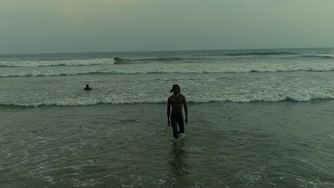 The artist walking out into the sea, back to camera, flat grey water to the horizon