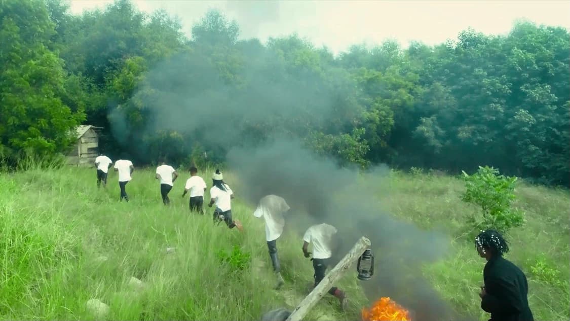 A line of figures in white running through long grass as coloured smoke drifts across the field