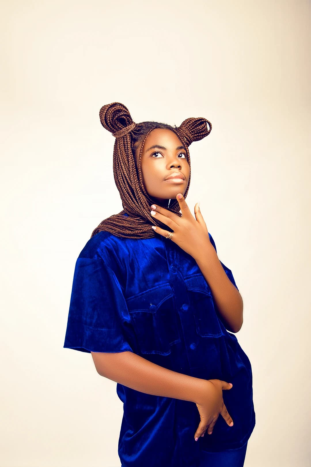 Braids drawn over the head like a hood, hand at the jaw, in an oversized blue velvet shirt