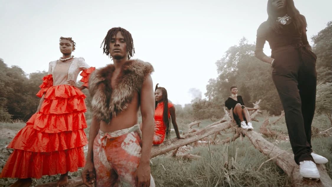 The full cast among fallen logs — red ruffled dresses, fur, and the artist centre frame