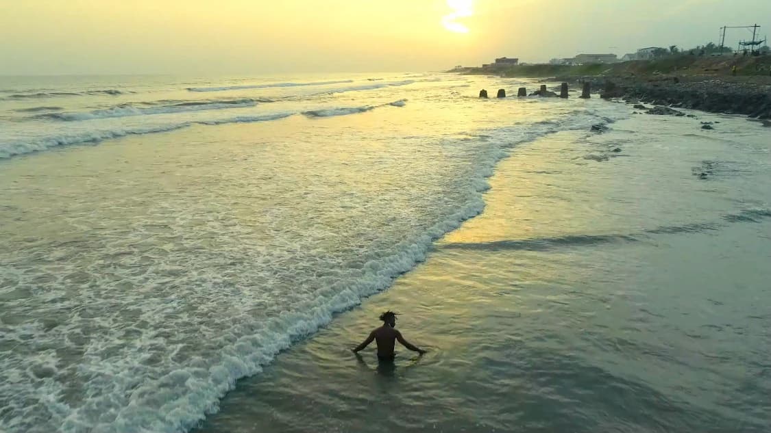 Wide of the shoreline at sunset, a lone figure standing waist-deep in the surf