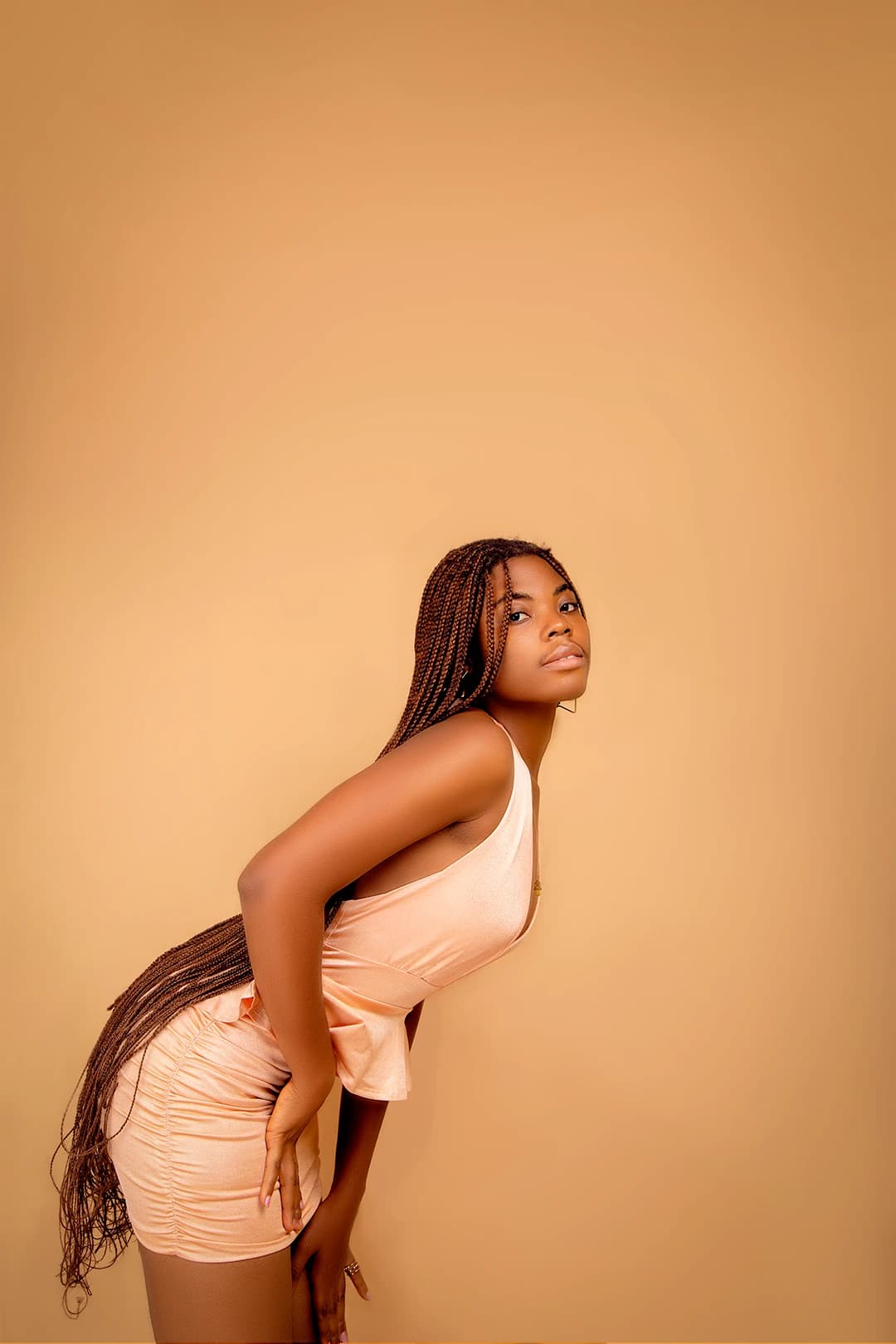 Bent forward in profile against the tan backdrop, braids falling to the floor, chin turned back to camera
