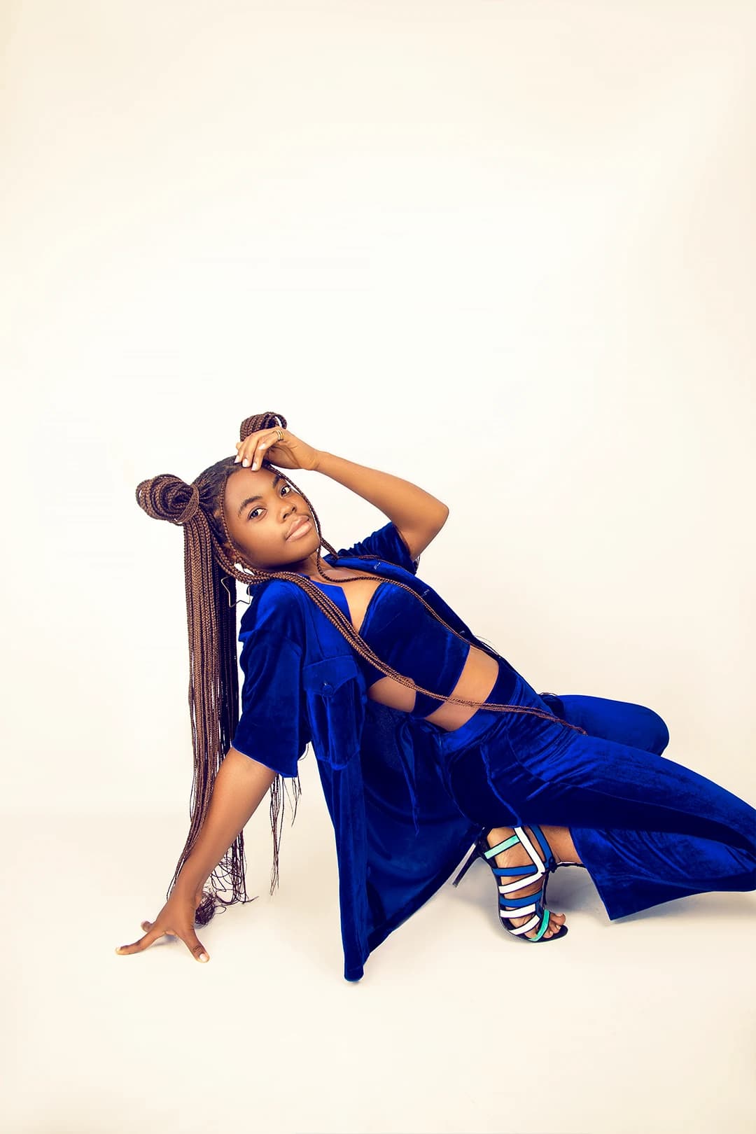 Seated on the floor in blue velvet and strappy heels, one hand to her hair, legs swept to the side