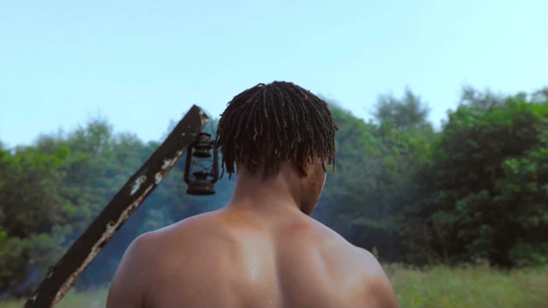 Shot from behind — bare shoulders and locs, the lantern and beam out of focus beyond