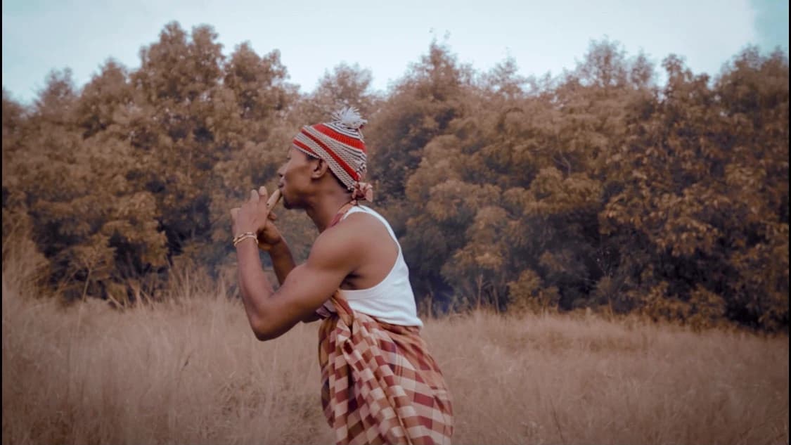 The artist in a headwrap and vest, hand raised to his face, in dry grass at the treeline