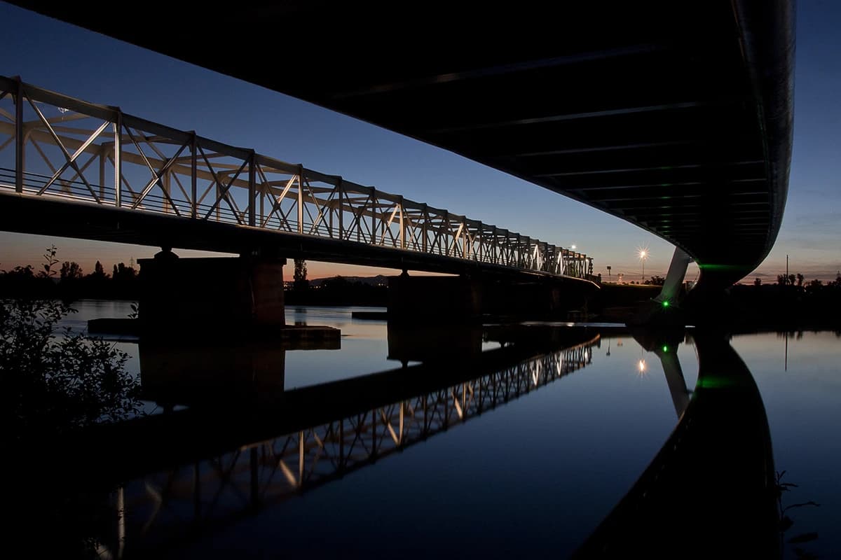 Looking along the underside of the road bridge at blue hour, the older truss bridge lit white beside it